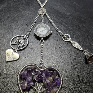 May include: A silver-toned pendant necklace featuring a small watch, a pentagram charm with a bird, a heart charm with "Blessed Be" text, and a heart-shaped pendant with a wire tree design filled with purple stones. The necklace has a chain and bell charm.