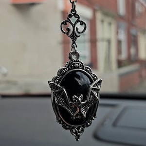 May include: A hanging ornament featuring a silver-toned metal frame with an ornate filigree design. The center contains a smooth, oval black stone with a silver bat figure mounted on top. A single black bead sits above the frame on a metal chain.