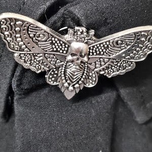 May include: A silver-toned metal brooch shaped like a moth with an intricate skull design on its thorax. The wings feature detailed embossed patterns including dots, lines, and geometric shapes. The brooch is pinned to a black fabric garment.