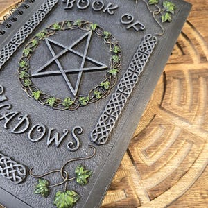 May include: A spiral-bound journal titled "BOOK OF SHADOWS" with a pentagram design. The cover is dark gray with Celtic knot and ivy leaf accents. The pentagram is encircled by a green ivy wreath.