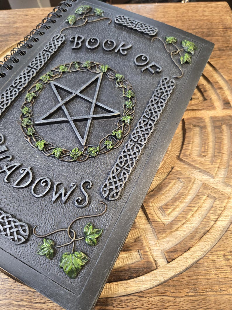 May include: A spiral-bound journal titled "BOOK OF SHADOWS" with a pentagram design. The cover is dark gray with Celtic knot and ivy leaf accents. The pentagram is encircled by a green ivy wreath.
