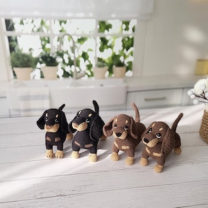 May include: Four crocheted dachshund dog toys. Two are black and brown, and two are brown and tan. The toys are sitting on a white wooden surface.