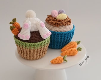 May include: Two crocheted Easter cupcakes, one with a white bunny and pink ears, the other with a brown nest and colorful eggs. Both cupcakes are decorated with green and brown frosting and are sitting on a white cake stand with three orange crocheted carrots.