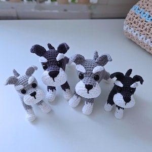 Könnte beinhalten: Vier handgefertigte Häkelfiguren von Hunden in verschiedenen Grau-, Schwarz- und Weißtönen. Jeder Hund hat ein schnauzerähnliches Aussehen mit weißer Schnauze, Pfoten und Augenbrauen. Die Hunde werden auf einer weißen Oberfläche präsentiert.