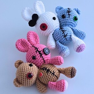 May include: A collection of handmade crochet stuffed animals in various colors. The toys include a white bunny with a black ear, a blue bear, a pink bunny, and two brown bears. Each toy has stitched details and button eyes.