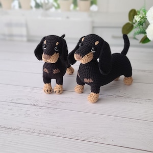 May include: Two black and brown crocheted dachshund dog plushies. The larger dachshund is standing with its legs outstretched, while the smaller dachshund is standing with its legs together.