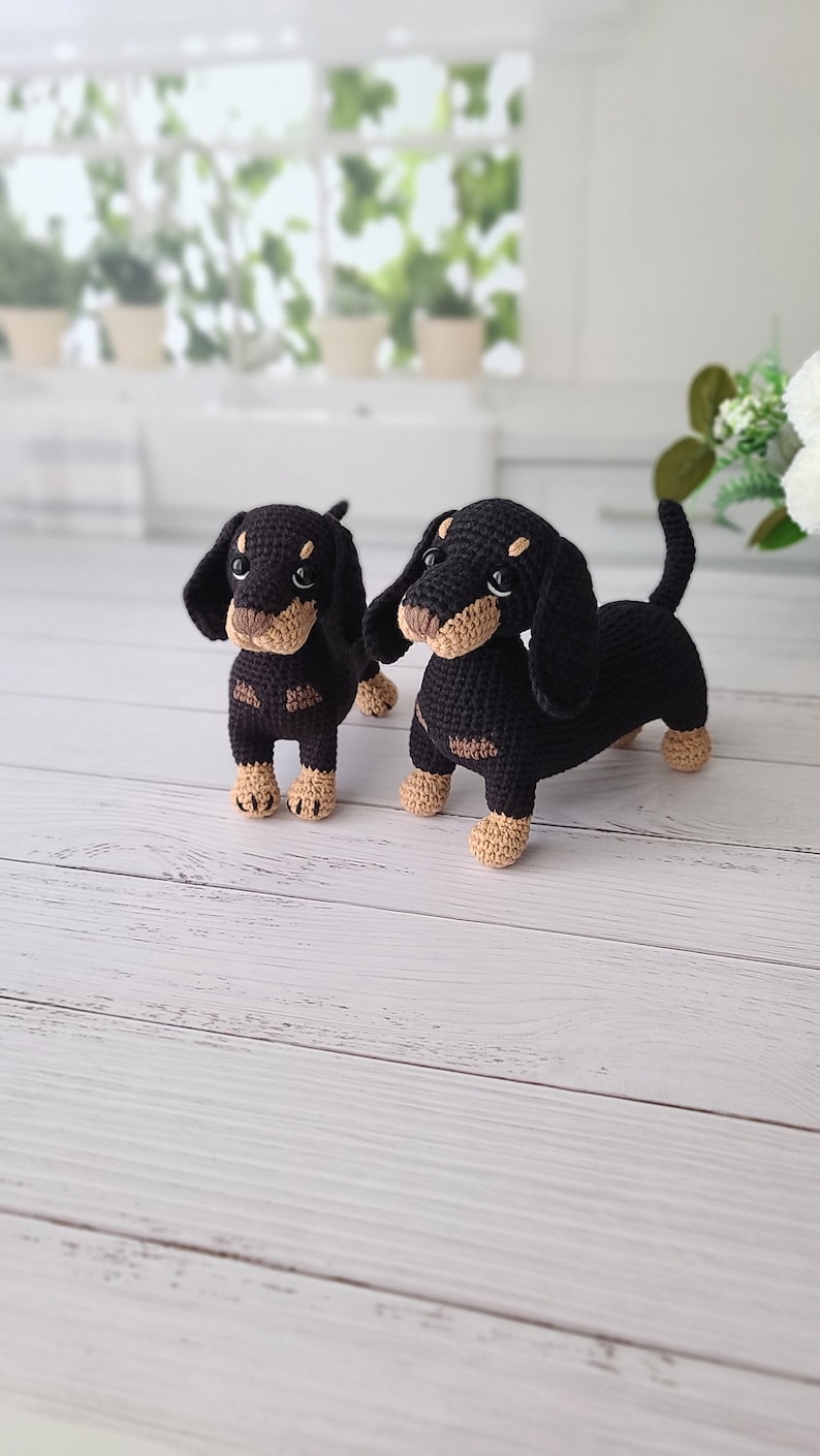 May include: Two black and brown crocheted dachshund dog plushies. The larger dachshund is standing with its legs outstretched, while the smaller dachshund is standing with its legs together.