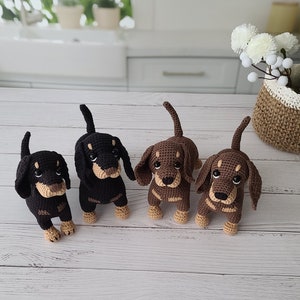 May include: Four crocheted dachshund dog toys. Two are brown and tan, and two are black and tan. The toys are sitting on a white wooden surface.
