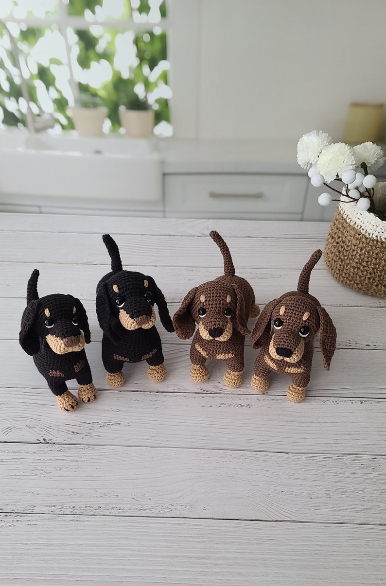 May include: Four crocheted dachshund dog toys. Two are brown and tan, and two are black and tan. The toys are sitting on a white wooden surface.