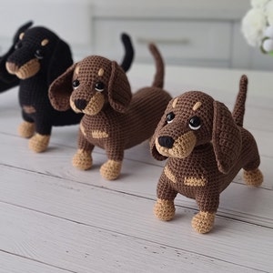 May include: Three crocheted dachshund dog toys. The dogs are brown, black, and brown with tan accents. The dogs are standing on a white wooden surface.