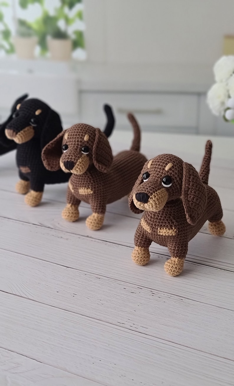 May include: Three crocheted dachshund dog toys. The dogs are brown, black, and brown with tan accents. The dogs are standing on a white wooden surface.