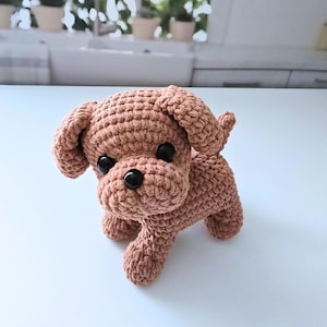 May include: A brown crocheted stuffed dog toy with black eyes. The dog is standing on a white surface.
