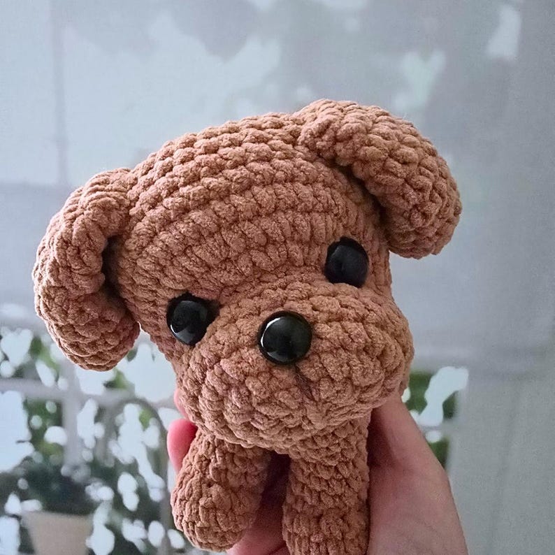 May include: A brown crocheted stuffed dog toy with black button eyes. The dog is sitting up with its front paws in the air.