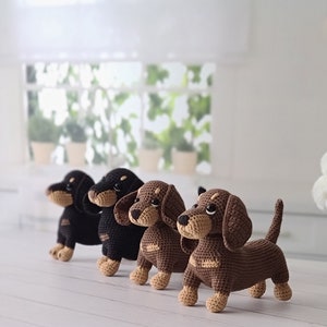 May include: Four crocheted dachshund dog toys in black, brown, and tan colors. The dogs are standing on a white wooden surface.