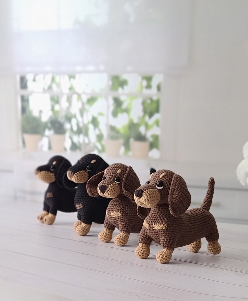 May include: Four crocheted dachshund dog toys in black, brown, and tan colors. The dogs are standing on a white wooden surface.