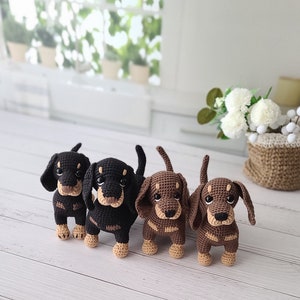 May include: Four crocheted dachshund dog plushies in black, brown, and tan colors. The plushies are standing on a white wooden surface.