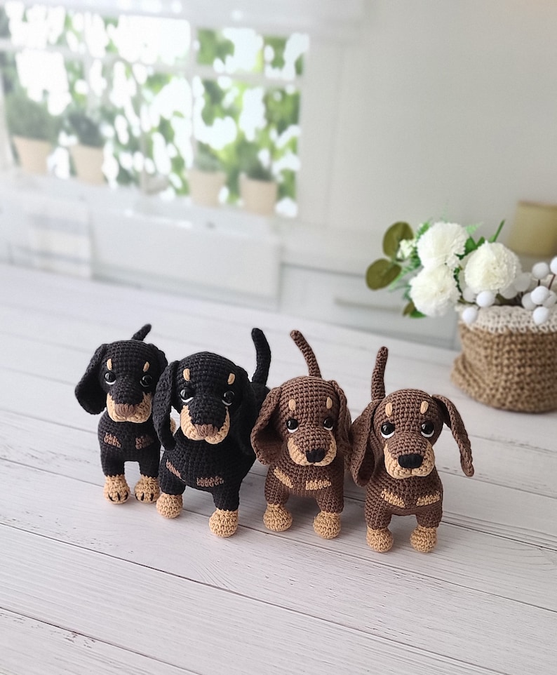 May include: Four crocheted dachshund dog plushies in black, brown, and tan colors. The plushies are standing on a white wooden surface.