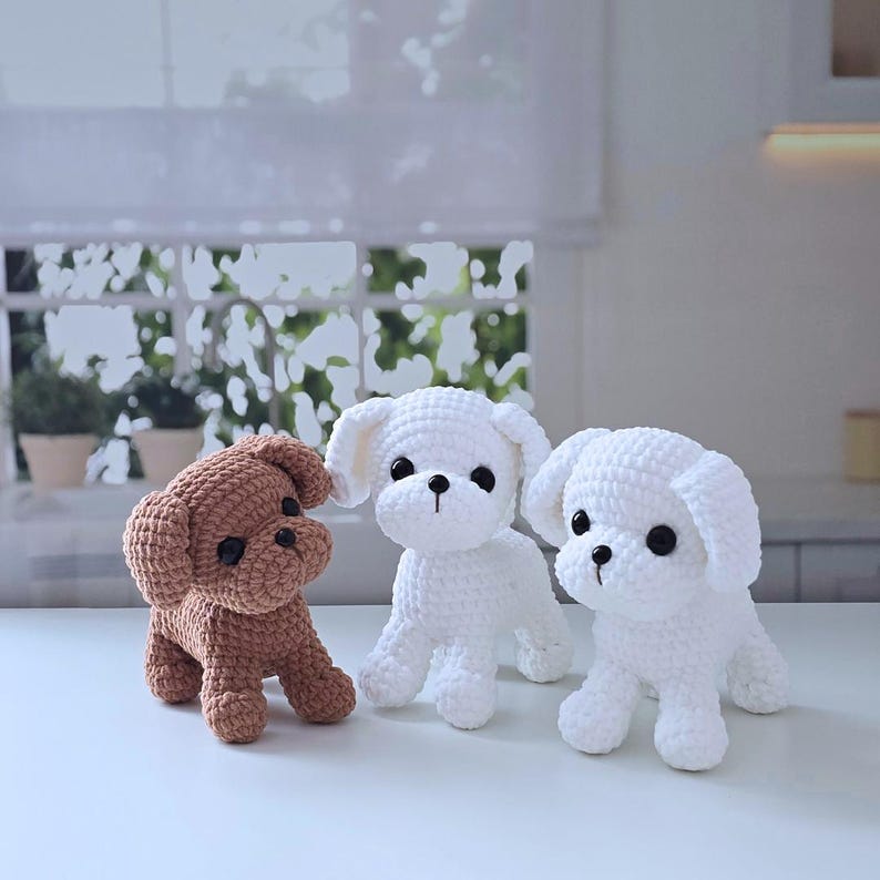 May include: Three crocheted stuffed dog toys. One brown dog, and two white dogs. The dogs are sitting on a white surface.