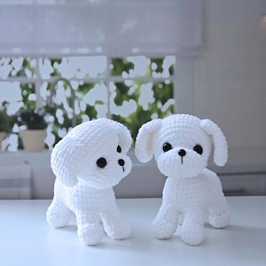 May include: Two white crocheted stuffed dogs with black eyes. The dogs are standing on a white surface.