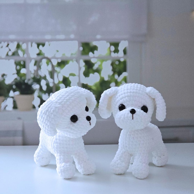 May include: Two white crocheted stuffed dogs with black eyes. The dogs are standing on a white surface.