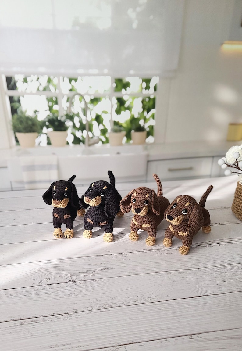 May include: Four crocheted dachshund dog toys. Two are black and brown, and two are brown and tan. The toys are sitting on a white wooden surface.