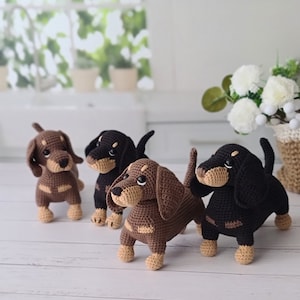 May include: Four crocheted dachshund dog plushies in brown, black, and tan colors. The plushies are standing on a white wooden surface.