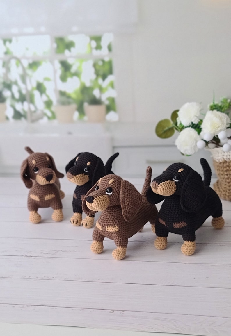 May include: Four crocheted dachshund dog plushies in brown, black, and tan colors. The plushies are standing on a white wooden surface.