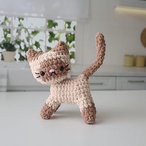 以下が含まれることがあります： 茶色とクリーム色の毛並みの、かぎ針編みで作った猫のぬいぐるみ。猫はピンクの鼻と黒い目を持っている。