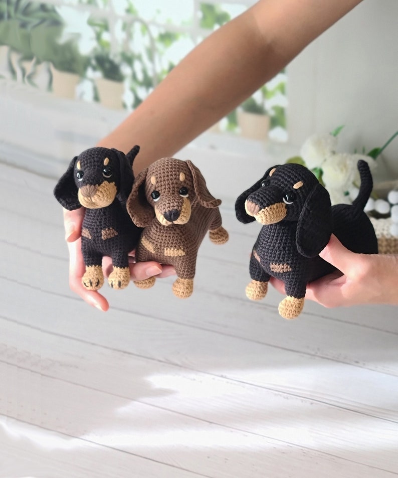 May include: Three crocheted dachshund dog plushies in black, brown, and tan colors. The plushies are sitting on a white wooden surface.
