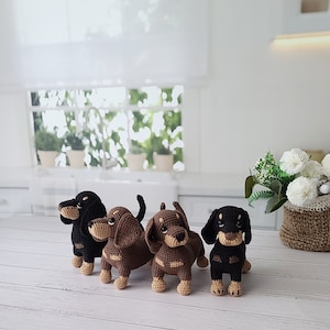 May include: Four crocheted dachshund dog toys in brown and black. The dogs are standing on a white wooden surface.