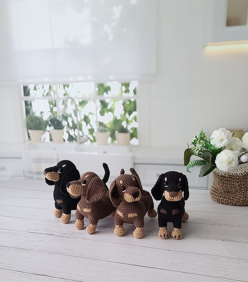 May include: Four crocheted dachshund dog toys in brown and black. The dogs are standing on a white wooden surface.