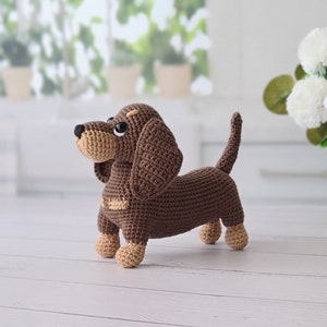 May include: A brown crocheted dachshund toy with brown eyes and a tan nose. The toy is standing on a white wooden surface.
