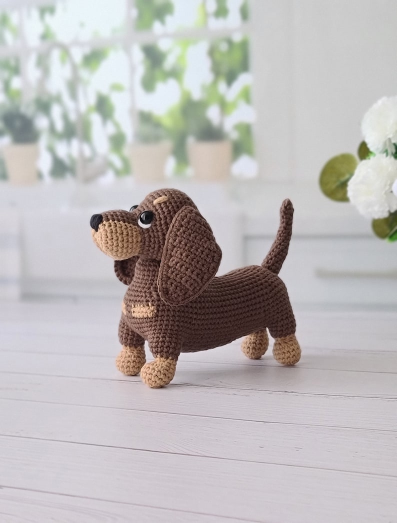 May include: A brown crocheted dachshund toy with brown eyes and a tan nose. The toy is standing on a white wooden surface.