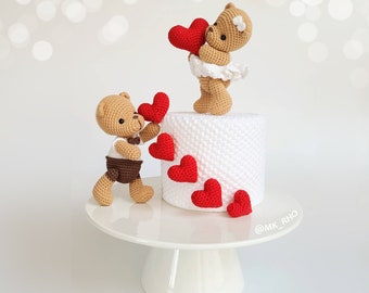 May include: Two crocheted teddy bears holding red hearts on top of a white cake with red hearts around the base. The cake is on a white cake stand. The text '@MK_RHO' is on the cake stand.