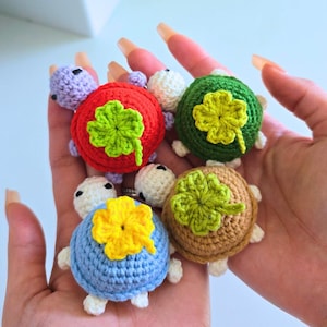 Può includere: Una collezione di cinque figurine di tartarughe all'uncinetto colorate. Ogni tartaruga ha un guscio di colore diverso: viola, rosso, verde, marrone e blu. Ogni guscio presenta un quadrifoglio o un motivo floreale. Le tartarughe hanno teste, piedi e code bianche.