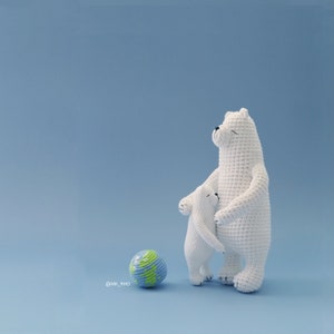 Pode incluir: Dois ursos polares de crochê branco em pé juntos, com um urso menor abraçando a cintura do maior. Um globo terrestre azul e verde fica na frente dos ursos sobre um fundo azul claro.
