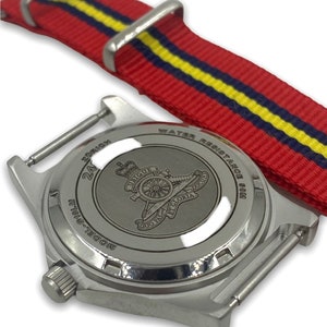 Puede incluir: Un reloj plateado con una correa de nailon a rayas rojas, amarillas y azules. La esfera del reloj no es visible, pero la parte posterior del reloj tiene un dise&ntilde;o grabado de un ca&ntilde;&oacute;n y las palabras "Ubique" y "Quo Fas Et Gloria Ducit".