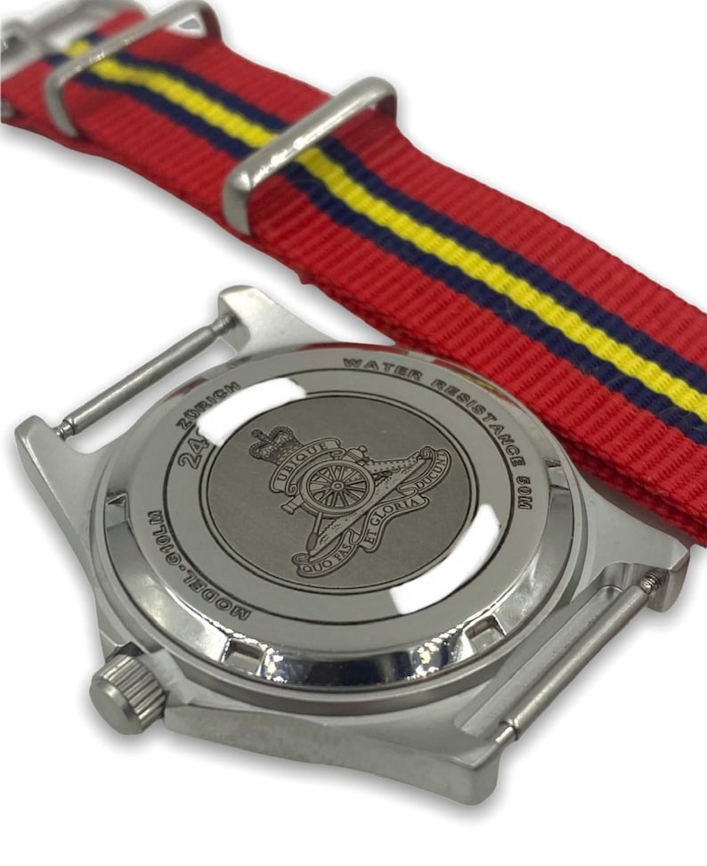 Puede incluir: Un reloj plateado con una correa de nailon a rayas rojas, amarillas y azules. La esfera del reloj no es visible, pero la parte posterior del reloj tiene un dise&ntilde;o grabado de un ca&ntilde;&oacute;n y las palabras "Ubique" y "Quo Fas Et Gloria Ducit".