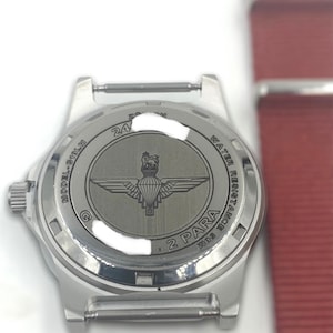Puede incluir: Reloj plateado con una correa de acero inoxidable. La parte posterior del reloj tiene un logotipo de 2 PARA y el texto "WATER RESISTANT 100M MODEL 6-101M 24".