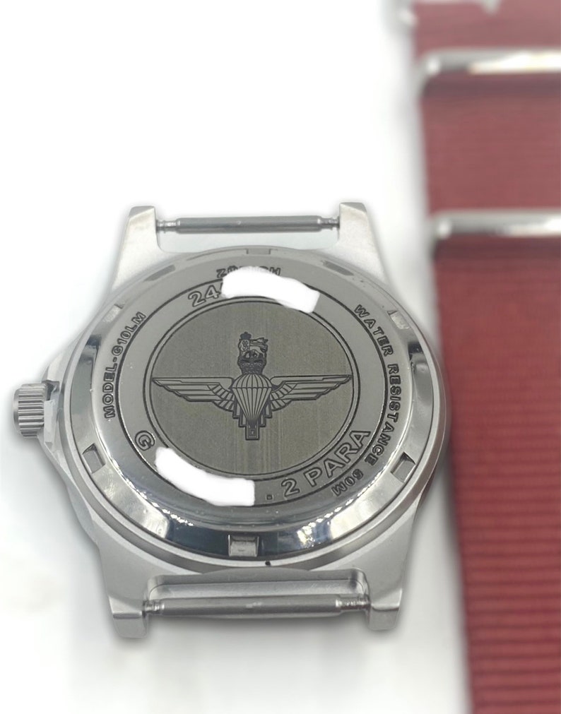 Puede incluir: Reloj plateado con una correa de acero inoxidable. La parte posterior del reloj tiene un logotipo de 2 PARA y el texto "WATER RESISTANT 100M MODEL 6-101M 24".