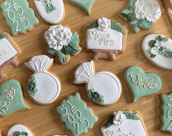 Puede incluir: Una colección de galletas de azúcar decoradas en una bandeja de madera. Las galletas presentan diseños temáticos de boda, incluyendo anillos de diamantes, corazones con el texto "We do" y letreros que dicen "Mr & Mrs". Las galletas están decoradas con glaseado blanco y verde.
