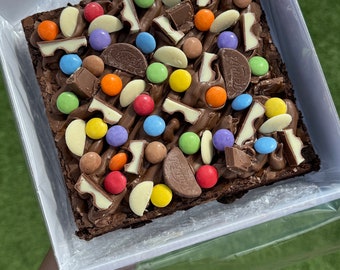 May include: A square chocolate brownie in a white box, topped with chocolate frosting, colorful candies, and chocolate pieces. The brownie is in a clear plastic container, held over green grass.