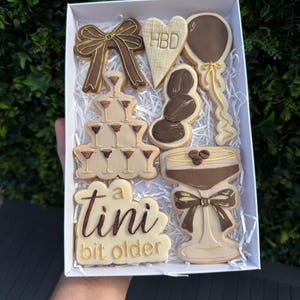 Op de afbeelding: Een doos met versierde koekjes met een verjaardagsthema. De koekjes hebben de vorm van een strik, champagneglazen, een ballon, koffiebonen en een martiniglas. De koekjes zijn bruin en beige met gouden accenten en de tekst "a tini bit older".