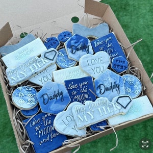 Peut inclure: Une boîte de biscuits décorés avec du glaçage bleu et blanc. Les biscuits ont diverses formes, notamment des cœurs et des hexagones, avec des messages tels que "Daddy" et "Love You". Certains biscuits présentent des motifs floraux.