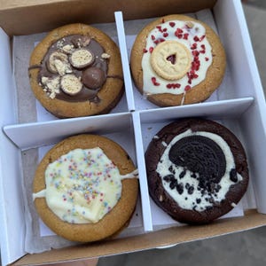 May include: A box of four gourmet cookies. One cookie has chocolate and candy pieces, another has a biscuit and red sprinkles, a third has colorful sprinkles, and the last has an Oreo cookie.