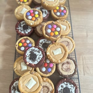 Op de afbeelding: Een assortiment aan gedecoreerde koekjes op een draadrek. De koekjes zijn bedekt met chocolade, sprinkles en witte chocolade. Sommige koekjes hebben een rode velvet basis.