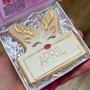Puede incluir: Una galleta de reno blanca y dorada con la nariz roja y la palabra "ABRIL" escrita en una pancarta. La galleta está en una caja blanca con papel triturado.