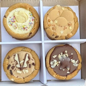 May include: Four gourmet cookies in a white box. Each cookie has a different topping: white chocolate rose with sprinkles, caramel with star-shaped candies, peanut butter with chocolate pieces, and chocolate with a bear-shaped candy.