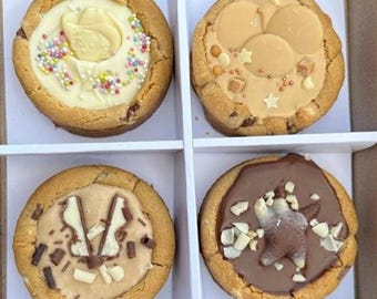 May include: Four gourmet cookies in a white box. Each cookie has a different topping: white chocolate rose with sprinkles, caramel with star-shaped candies, peanut butter with chocolate pieces, and chocolate with a bear-shaped candy.