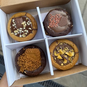 May include: A box of four gourmet cookies, each with a unique topping. The cookies are in a white box with dividers. Toppings include chocolate, nuts, and honeycomb. Each cookie is in its own compartment.
